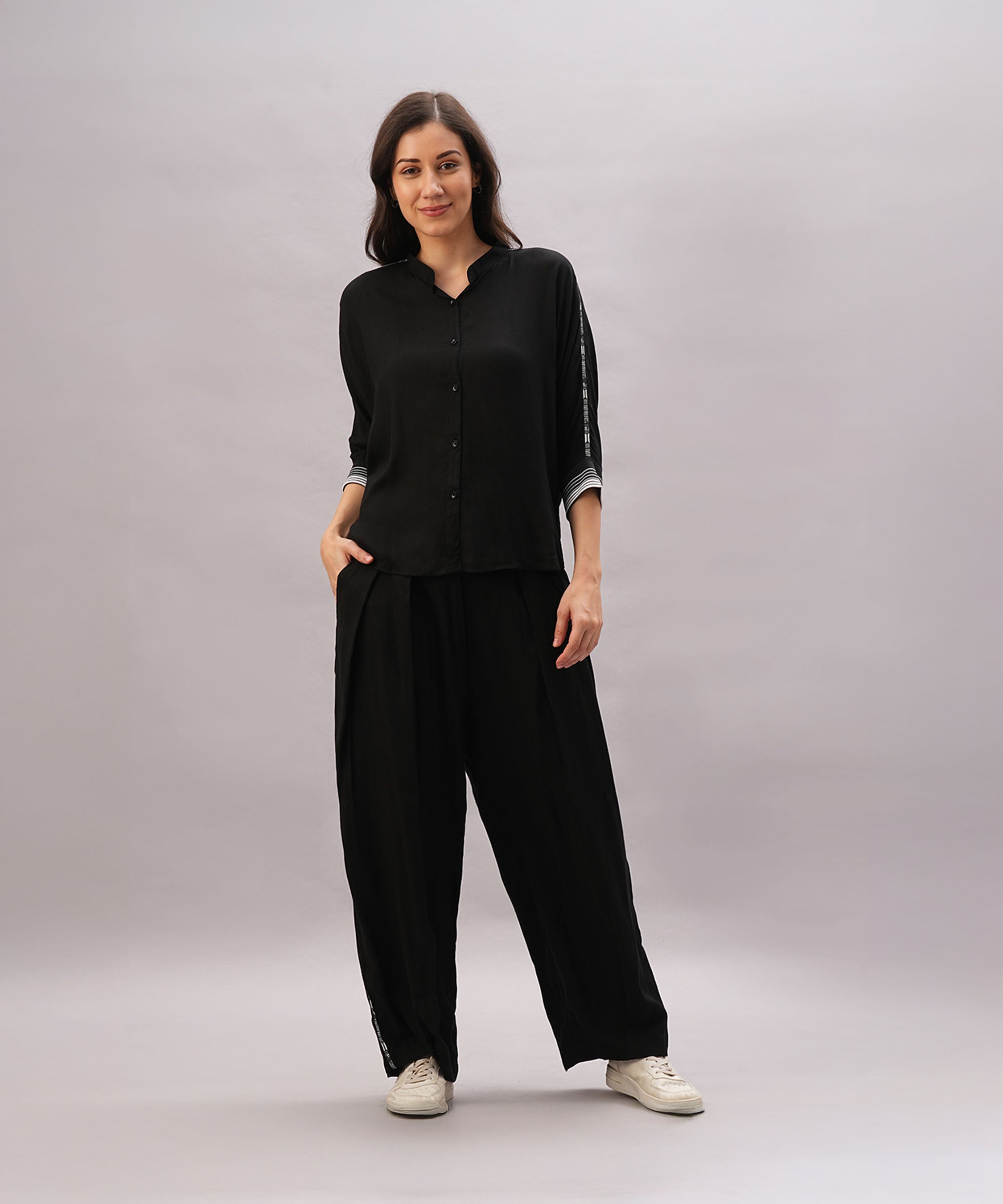 Woman in a Black Rayon Co-Ord Set featuring a chic top and relaxed pants, paired with white sneakers, stands with one hand in her pocket against a plain background.