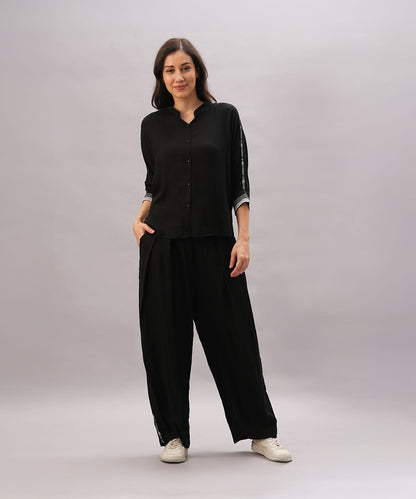 Woman in a Black Rayon Co-Ord Set featuring a chic top and relaxed pants, paired with white sneakers, stands with one hand in her pocket against a plain background.