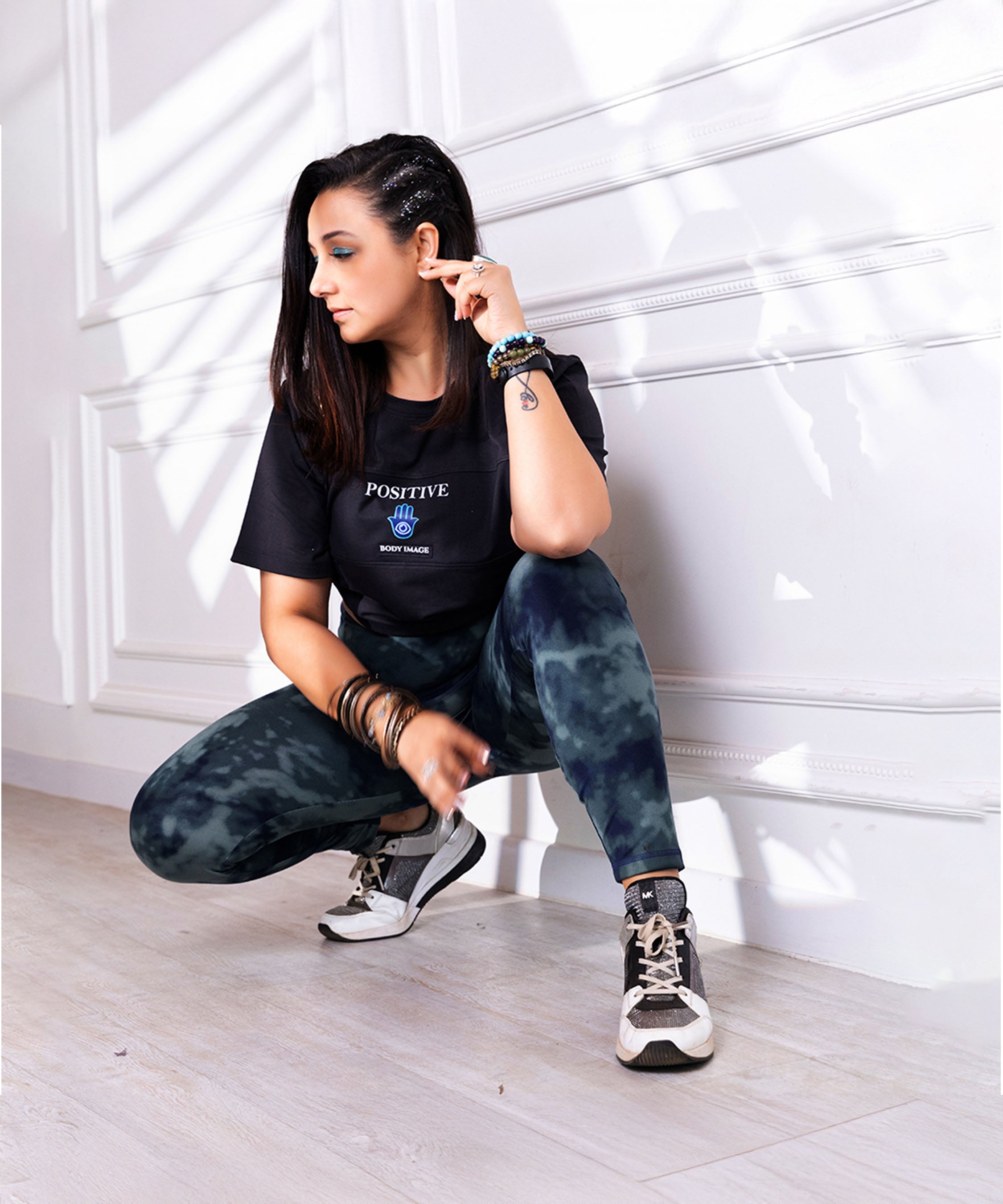 A woman in casual wear squats indoors by a sunlit white wall, wearing the Positive Body Image T Shirt, a black basic tee that celebrates confidence and self-love.