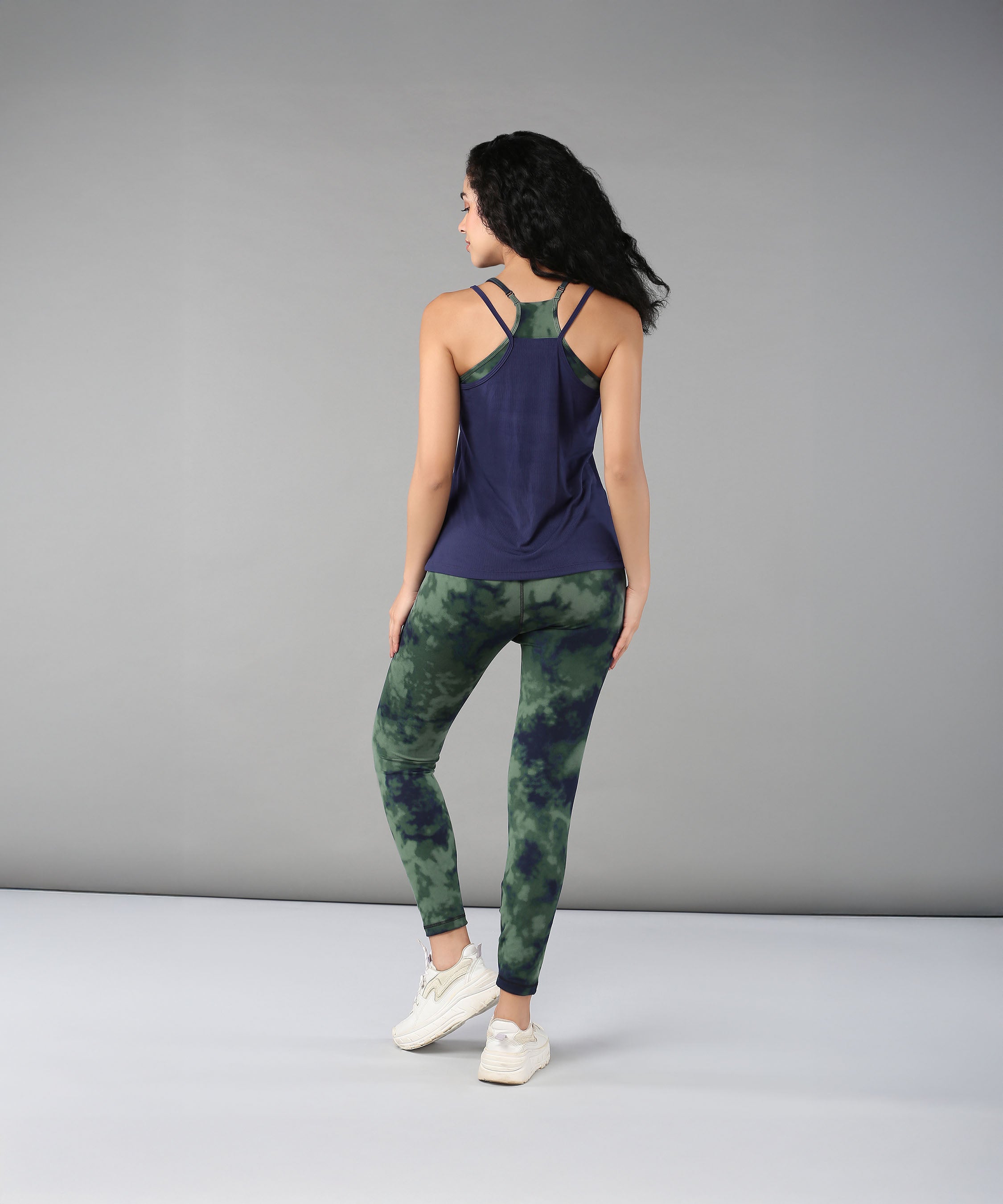 A woman faces away on a plain grey background, wearing the Tie & Dye Bra T shirt in green over a navy tank top.