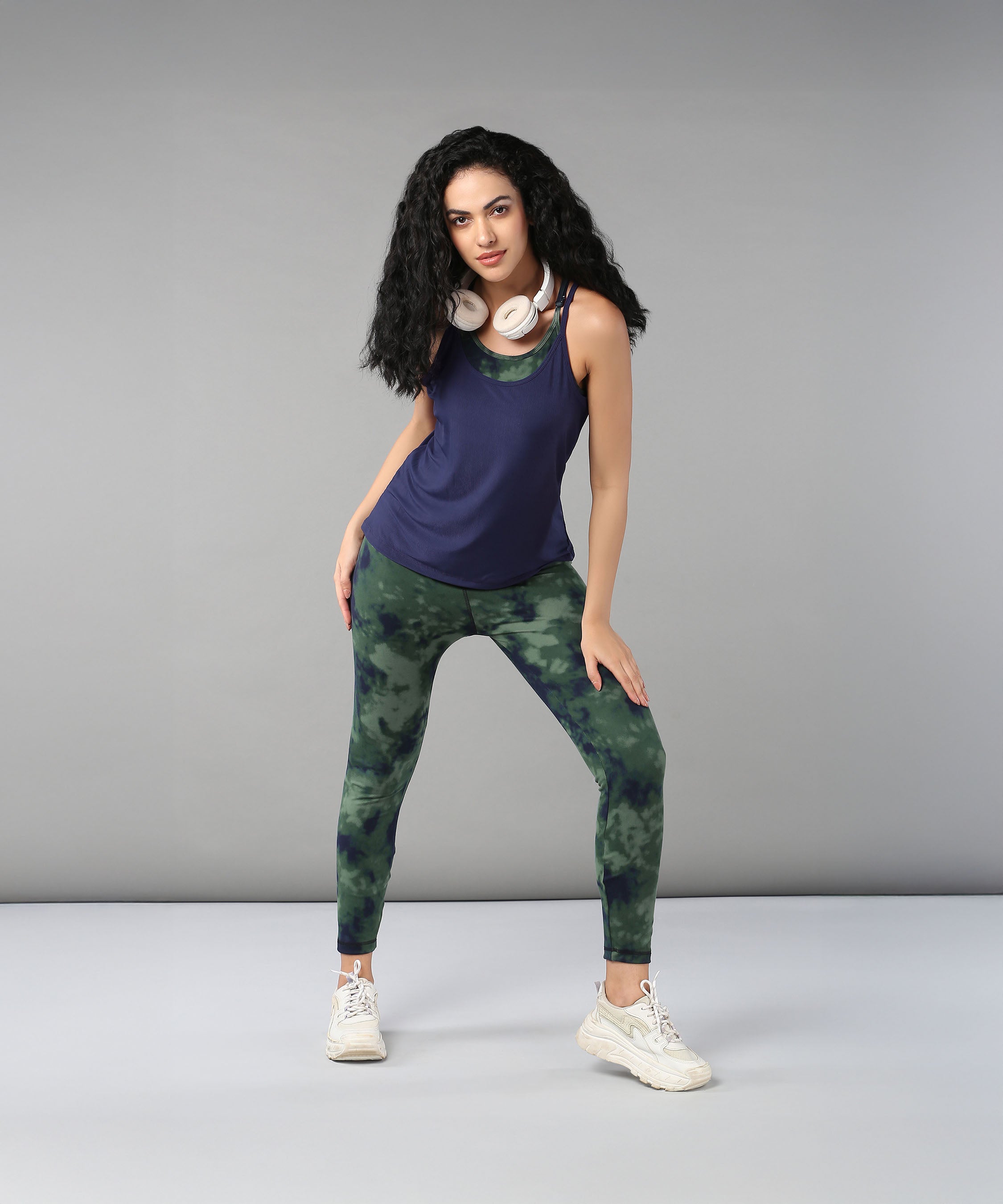 A woman with curly hair wears headphones, a Tie & Dye Bra T shirt, green leggings, and white sneakers as she poses confidently.