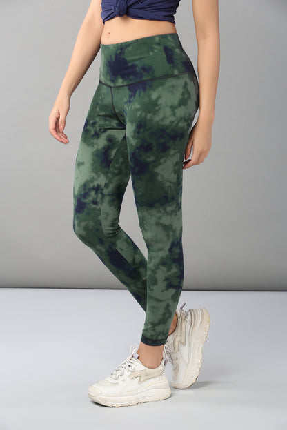 A woman wears Tie & Dye Leggings made from stretchy green fabric, paired with a blue top and white sneakers, standing against a plain background—a chic choice for any workout wardrobe.
