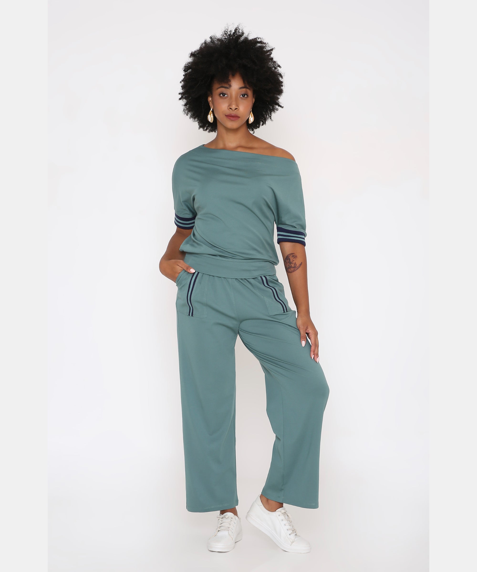 A woman wears the Sporty Sage Green Offshoulder Tracksuit Set, crafted from a soft Tencel-Spandex blend, styled with white sneakers against a plain white background.