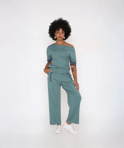 A woman with curly hair wears the Sporty Sage Green Off-shoulder Tracksuit Set, posing against a white background.
