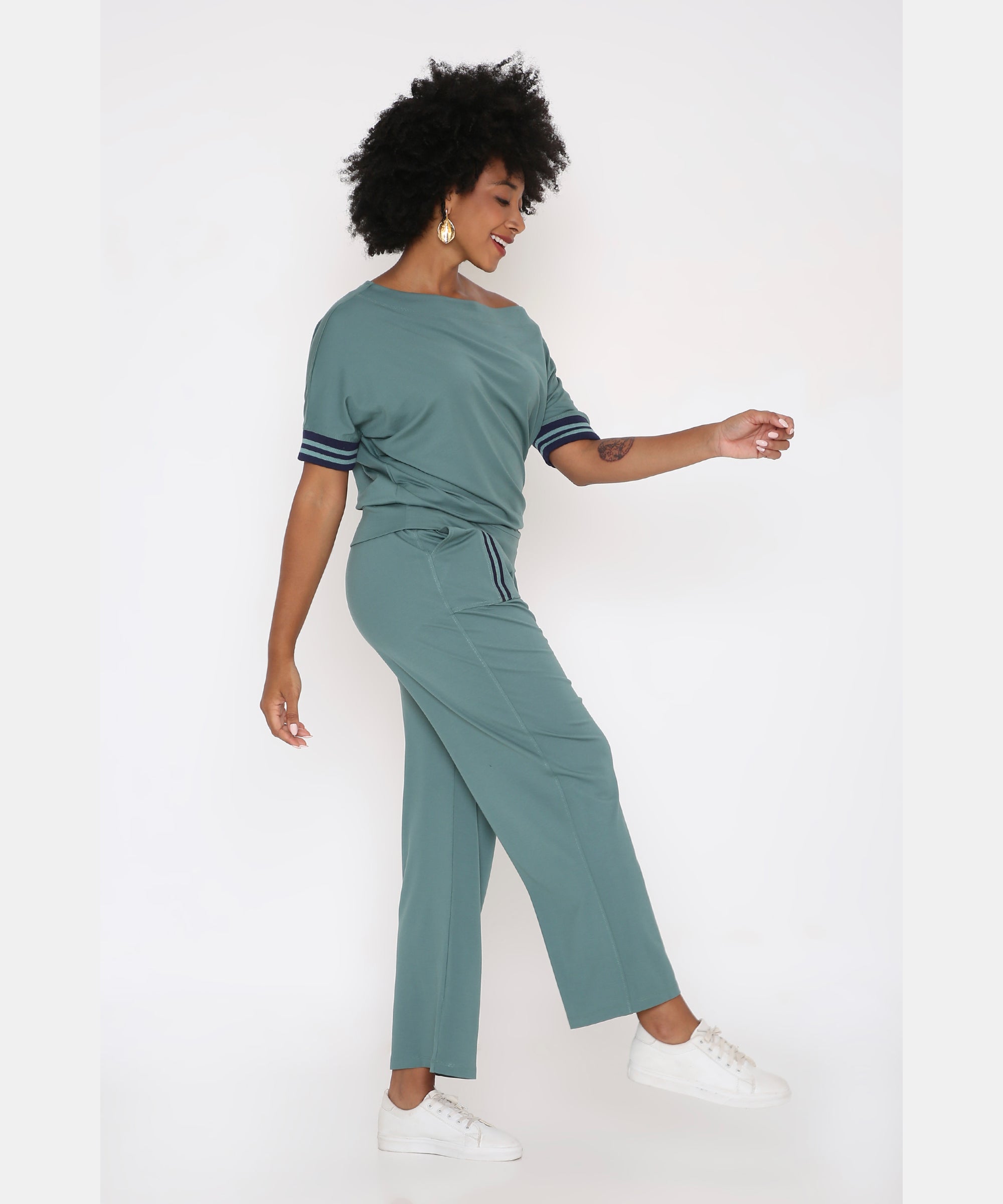 A woman in the Sporty Sage Green Offshoulder Tracksuit Set and white sneakers poses sideways against a white background, smiling.
