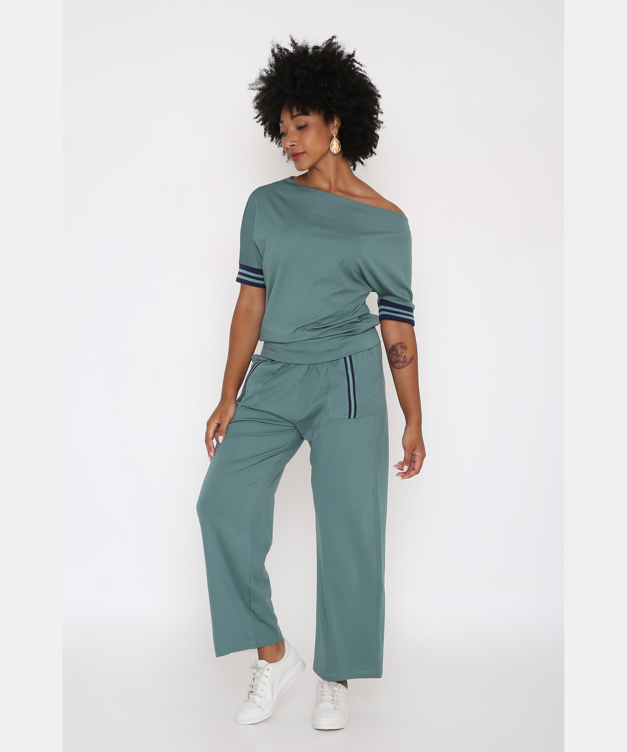 A woman wears the Sporty Sage Green Offshoulder Tracksuit Set, crafted from a soft Tencel-Spandex blend, styled with white sneakers against a white background.