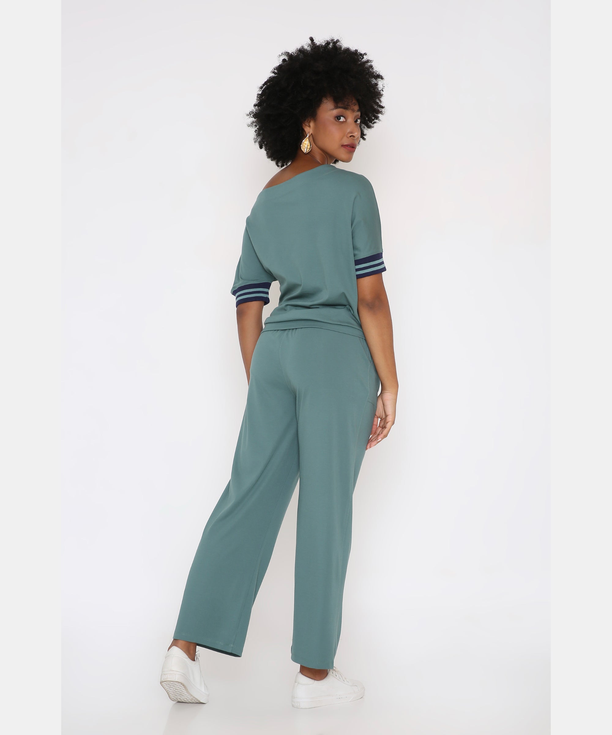 A woman with curly hair models the Sporty Sage Green Offshoulder Tracksuit Set with striped sleeves, looking over her shoulder against a white background.