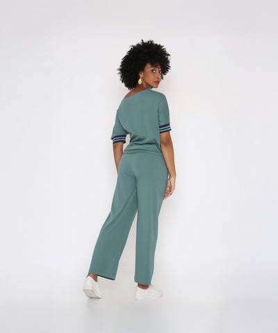 Woman with curly hair wearing the Sporty Sage Green Off-shoulder Tracksuit Set and white sneakers, standing and looking over her shoulder against a white background.
