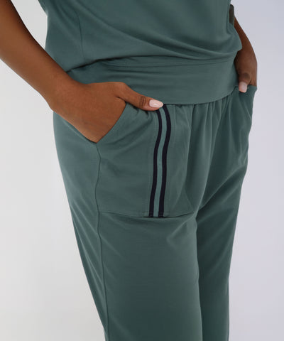 Person wearing the Sporty Sage Green Off-shoulder Tracksuit Set—sage green jogger pants with black double pocket stripes and hands in pockets, a stylish addition to any women's co-ord wardrobe.