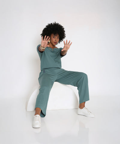 A woman wears the Sporty Sage Green Off-shoulder Tracksuit Set, sitting on a white arch and reaching her hands forward with a serious expression.