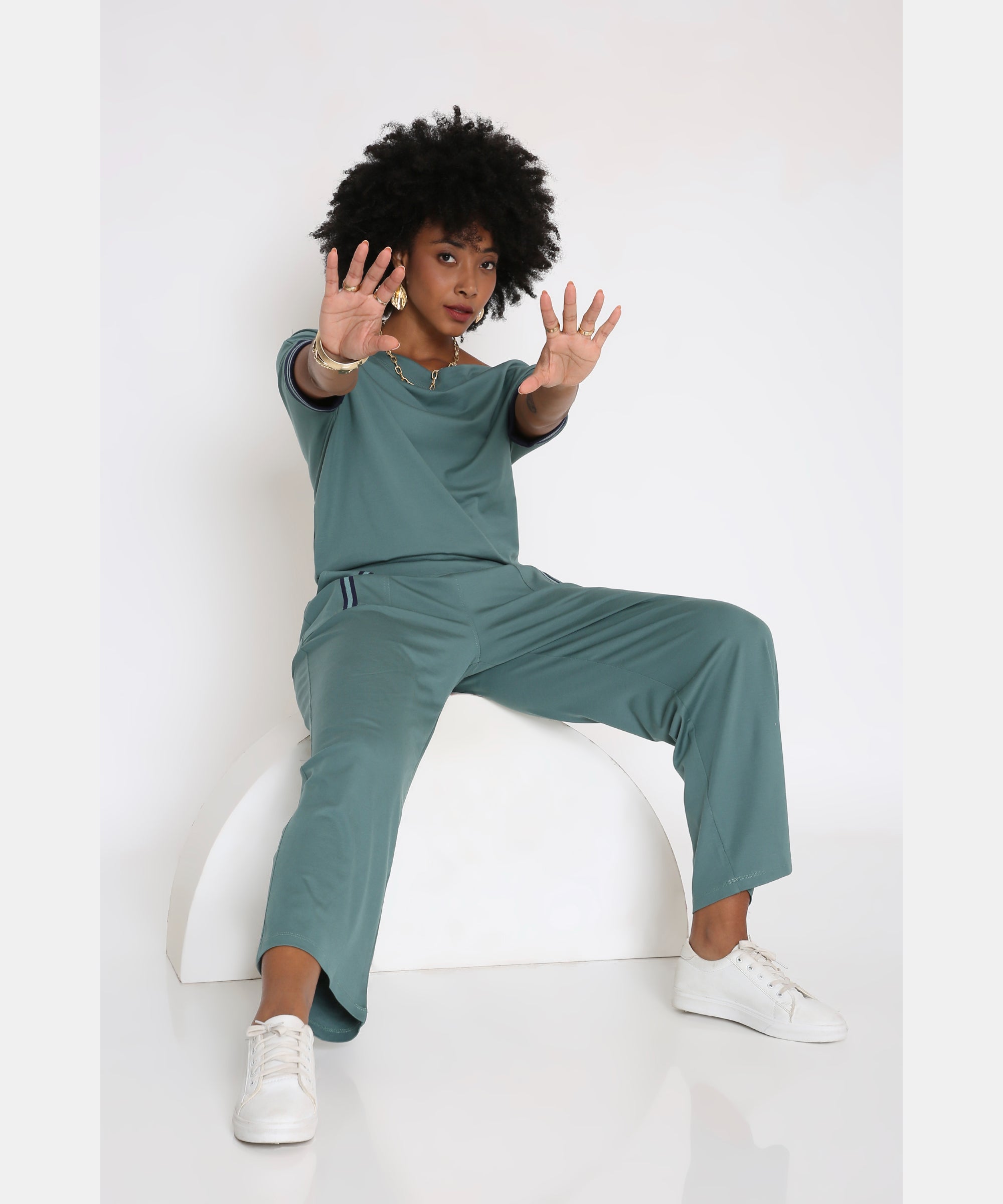 A woman wears the Sporty Sage Green Offshoulder Tracksuit Set, sitting on a white curved block and reaching forward with both hands while sporting white sneakers.