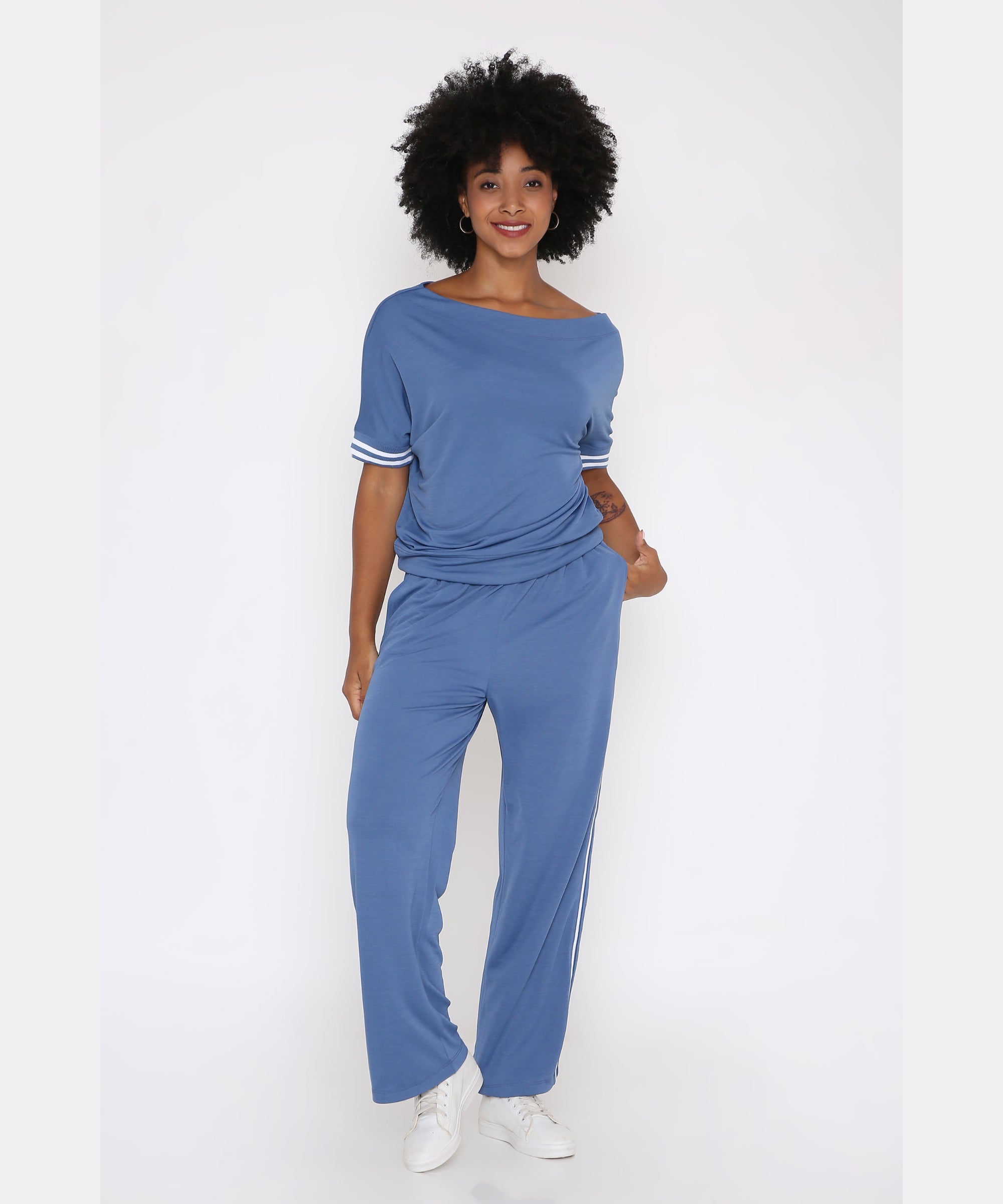 Woman in a Sporty Atlantic Blue Off-Shoulder Tracksuit Set with white sneakers, standing and smiling against a white background.