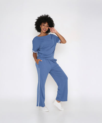 Smiling, a woman touches her hair while posing in the Sporty Atlantic Blue Off-Shoulder Tracksuit Set with white sneakers—a chic choice for an effortless athleisure vibe.