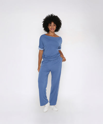 Woman with curly hair wears the Sporty Atlantic Blue Off-Shoulder Tracksuit Set, crafted from a soft Tencel-Spandex blend, paired with white sneakers, standing against a plain white background.