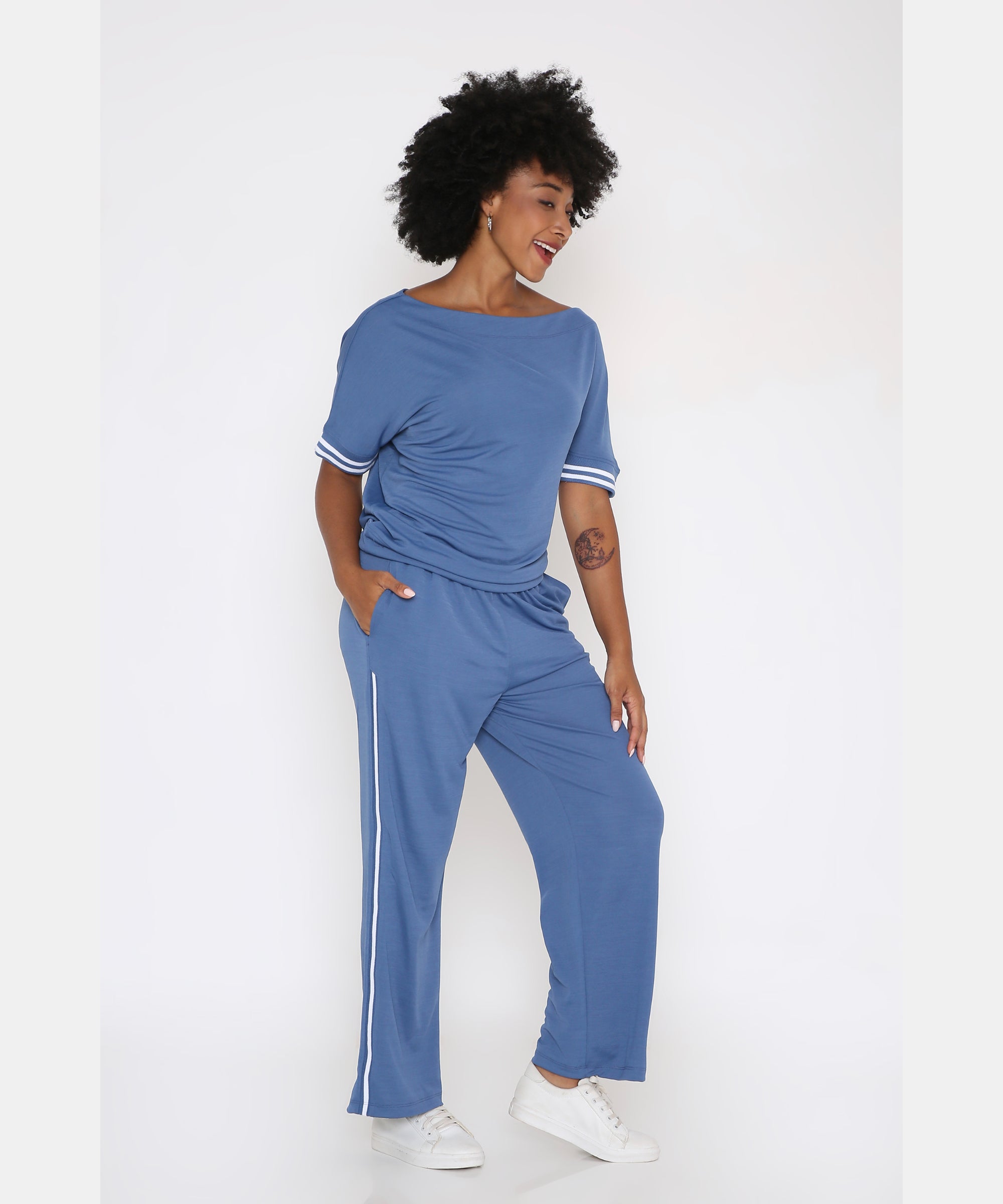 Woman smiles with hands in pockets, wearing the Sporty Atlantic Blue Off-Shoulder Tracksuit Set featuring white stripes, styled with white sneakers.