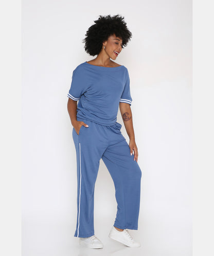 Woman smiles with hands in pockets, wearing the Sporty Atlantic Blue Off-Shoulder Tracksuit Set featuring white stripes, styled with white sneakers.