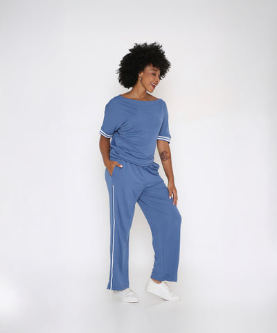 A woman wearing the Sporty Atlantic Blue Off-Shoulder Tracksuit Set and white sneakers smiles in a sporty athleisure look against a plain white background.