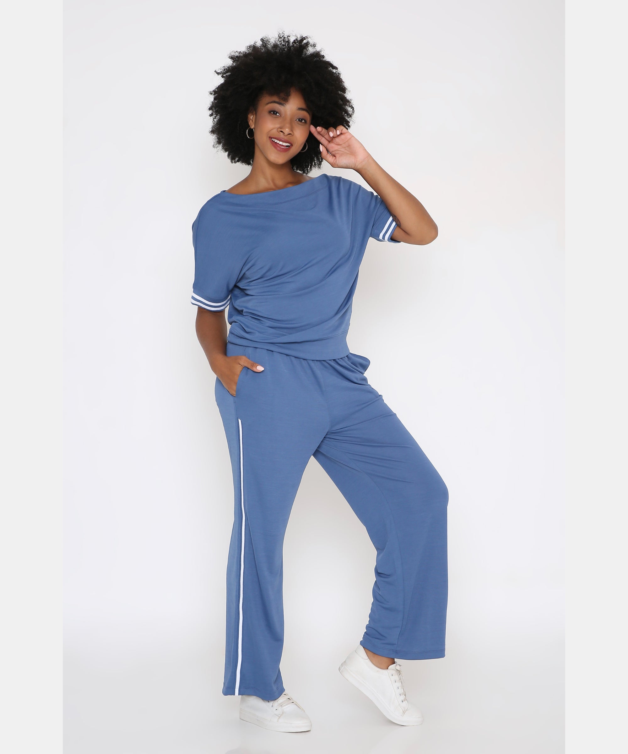 Woman smiling in the Sporty Atlantic Blue Off-Shoulder Tracksuit Set—Tencel-Spandex blend loungewear with white trim, styled with white sneakers and embracing athleisure chic against a white background.
