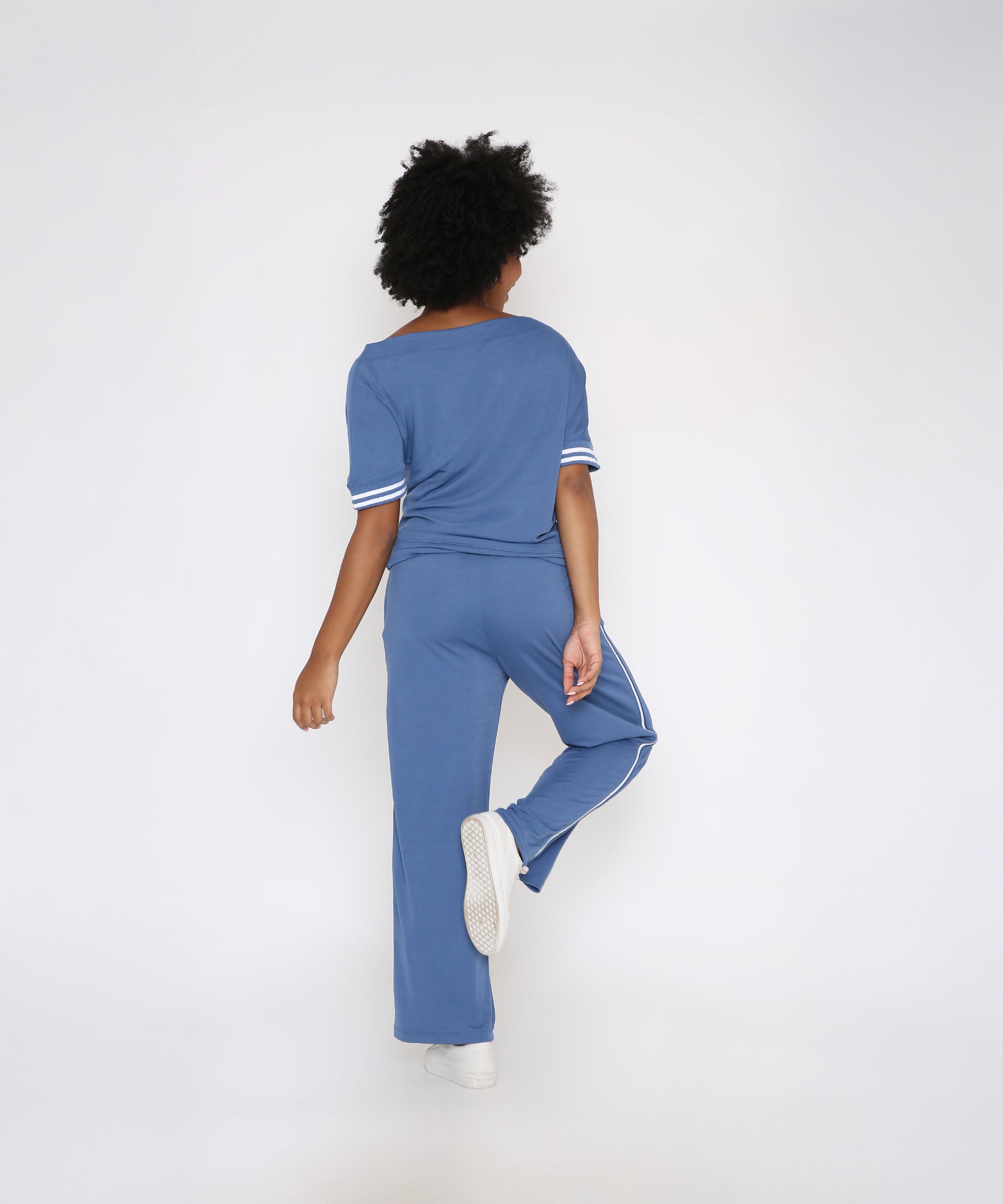 Wearing the Sporty Atlantic Blue Off-Shoulder Tracksuit Set and white sneakers, a person stands with their back to the camera, lifting one leg slightly.