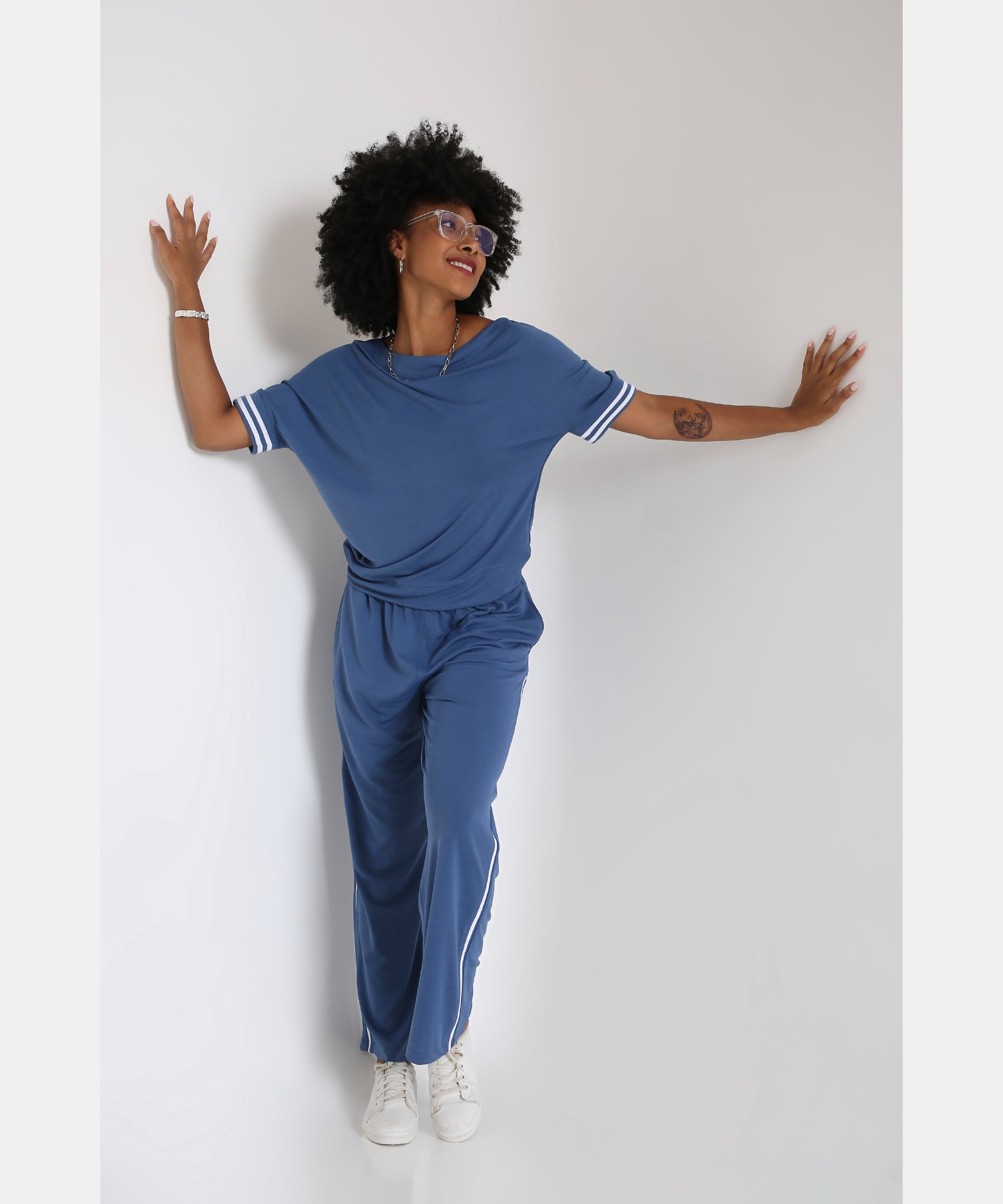 A woman smiles with arms outstretched, posing in the Sporty Atlantic Blue Off-Shoulder Tracksuit Set and white sneakers against a white wall.
