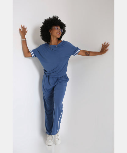 A woman smiles with arms outstretched, posing in the Sporty Atlantic Blue Off-Shoulder Tracksuit Set and white sneakers against a white wall.