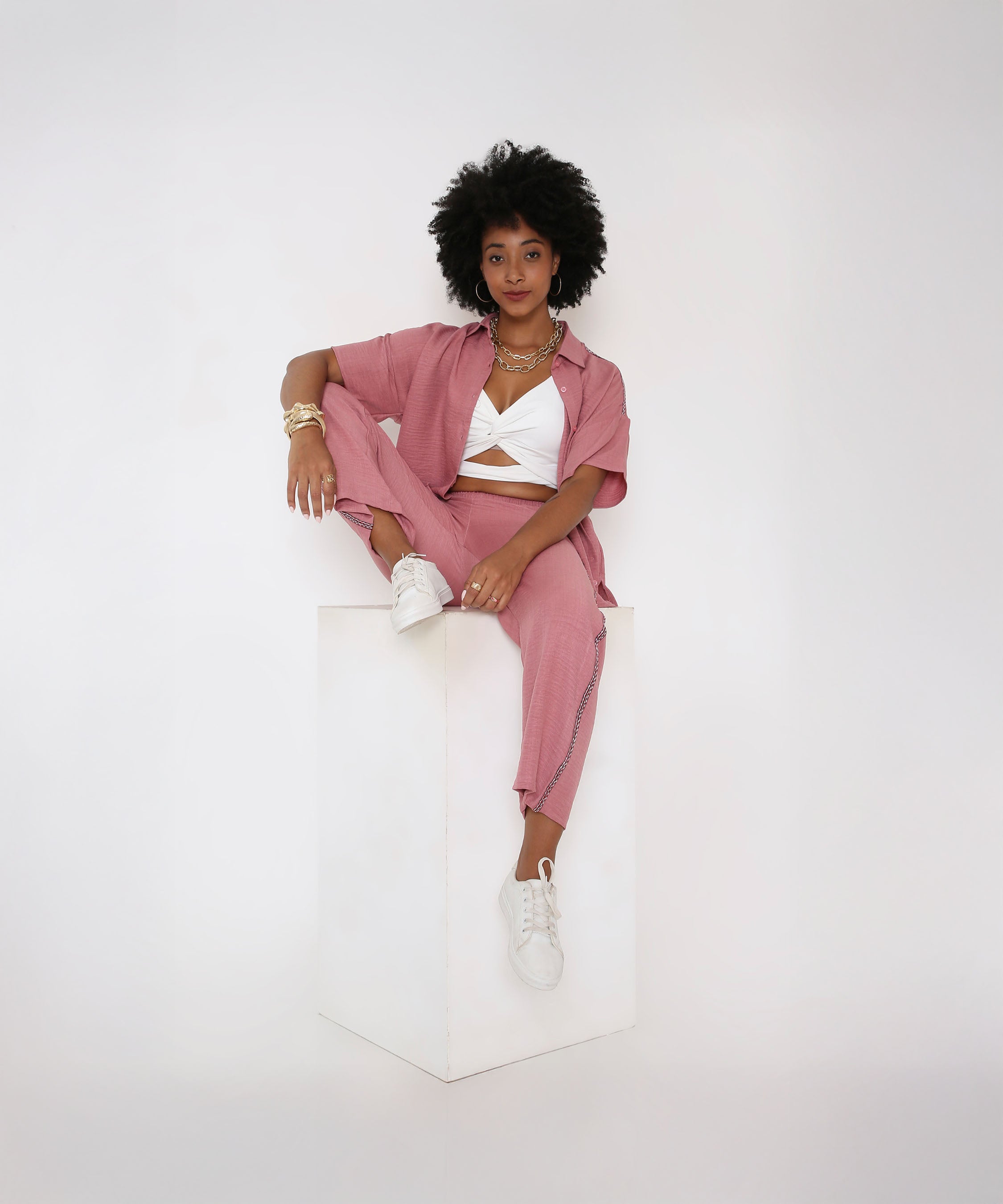Woman with natural hair sits on a white cube, wearing the Vintage Pink Loose Shirt and Pant Cord Set—this 2-piece set pairs perfectly with a white top and matching sneakers.