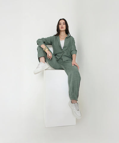 A woman in the Moss Green Breezy Cord Set and white sneakers sits confidently on a white cube against a plain background, highlighting the relaxed fit and modern style of this loungewear.