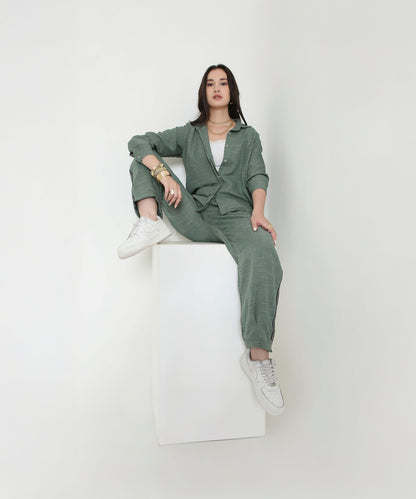 A woman in the Moss Green Breezy Cord Set and white sneakers sits confidently on a white cube against a plain background, highlighting the relaxed fit and modern style of this loungewear.