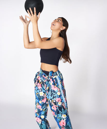Woman in floral pants and a Navy Blue Crop Top tosses a black ball upward, gazing at it against a plain background.