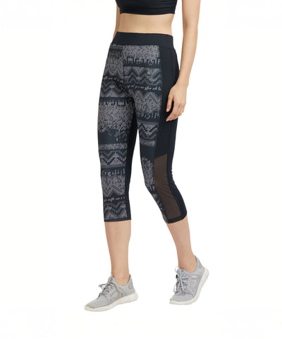 A woman wears the Printed Capri—patterned black capri-length track pants in soft cotton-spandex—with a black crop top and gray sneakers, set against a white background.