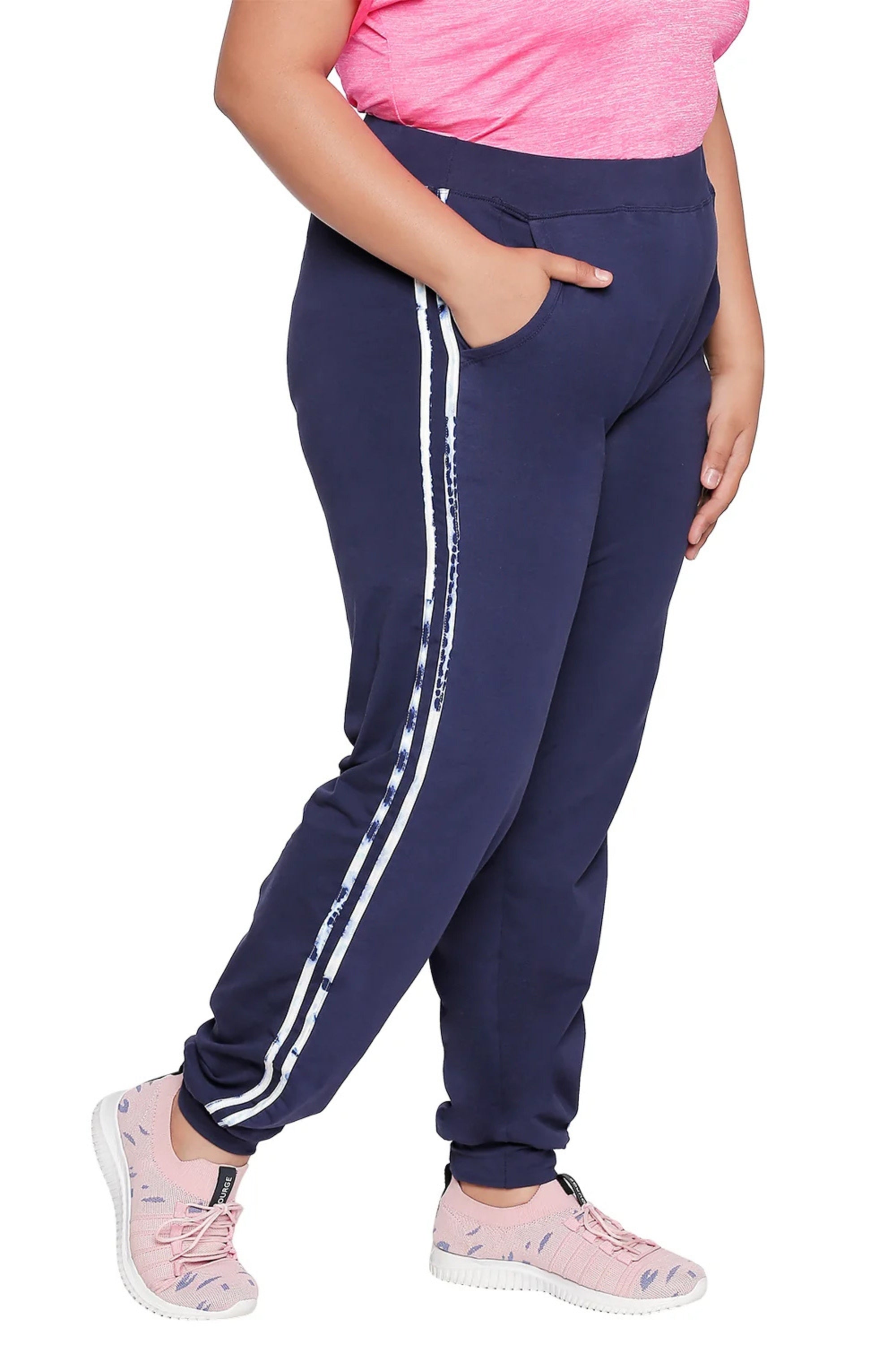 Wearing Joggers With Side Stripe - Plus Size in navy blue with white side stripes, paired with a pink top and sneakers. The high-stretch fabric offers comfort and style for any occasion. Model stands sideways.