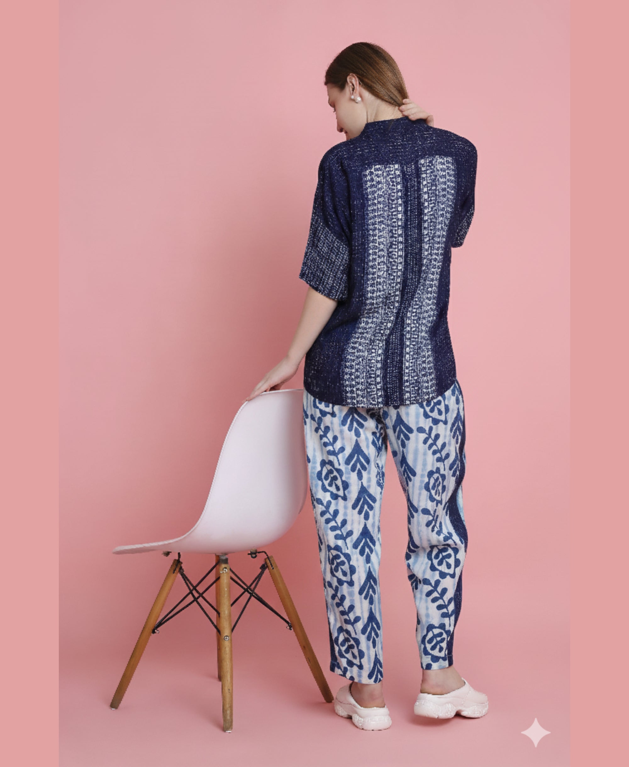 Woman wears the Indigo Printed Top with Tie Dye Printed Pant Lounge Set, facing away and resting her hand on a white chair against a pink background.
