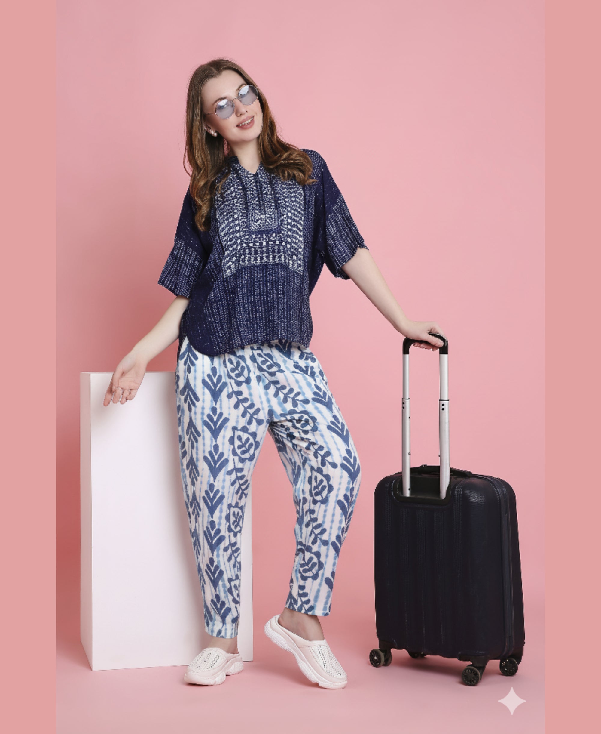 Woman in sunglasses wears the Indigo Printed Top with Tie Dye Printed Pant Lounge Set, standing with a suitcase against a pink background and smiling.
