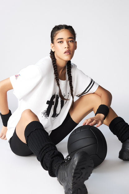 A young woman with braids sits on the floor, confidently holding a black basketball while wearing the 100% cotton Oversize Fly Tee featuring a sporty butterfly print.