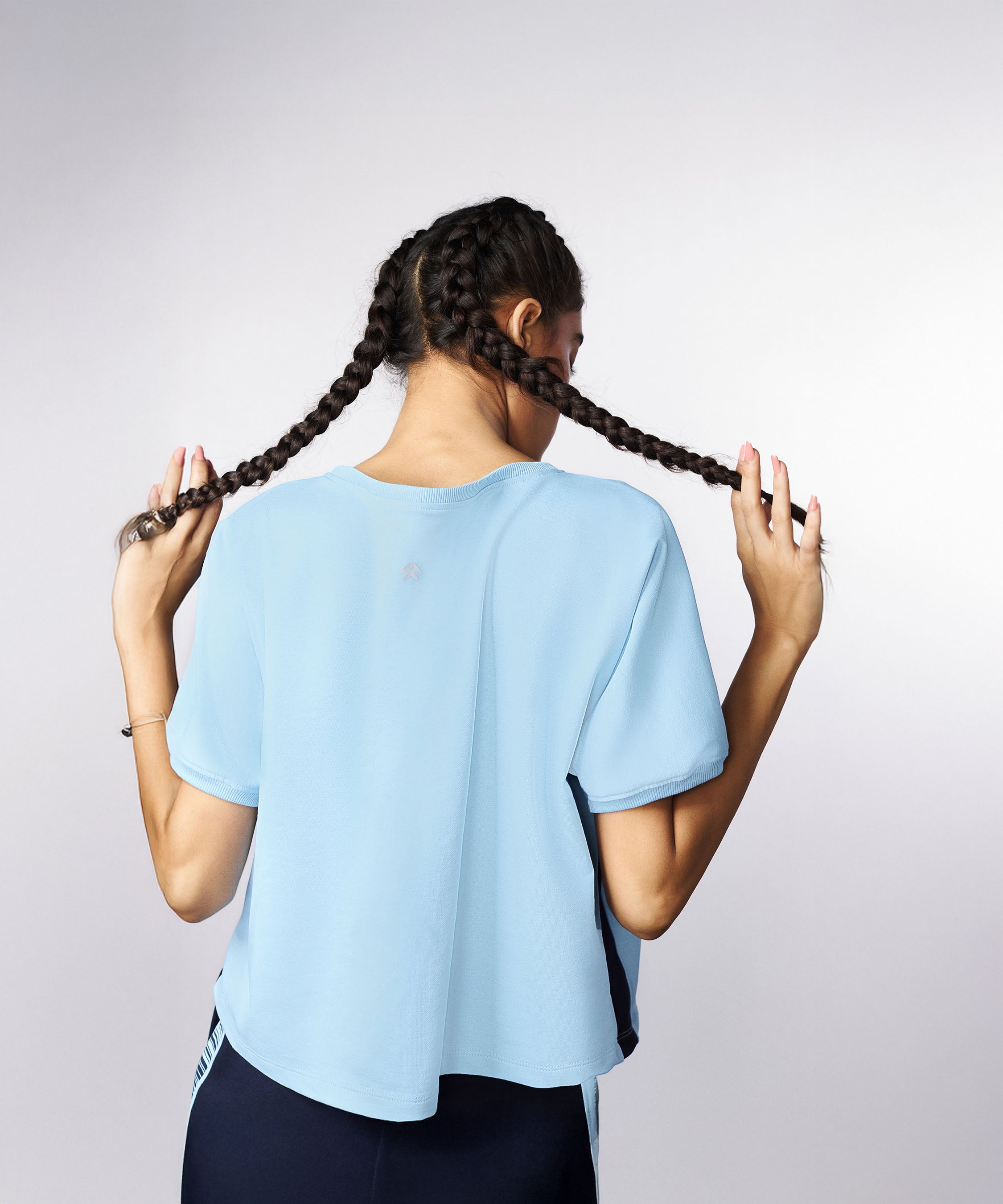 A woman with two braids, seen from behind, wears a light blue shirt and Tuna Active Barcode Printed Blue Shorts, holding her braids.