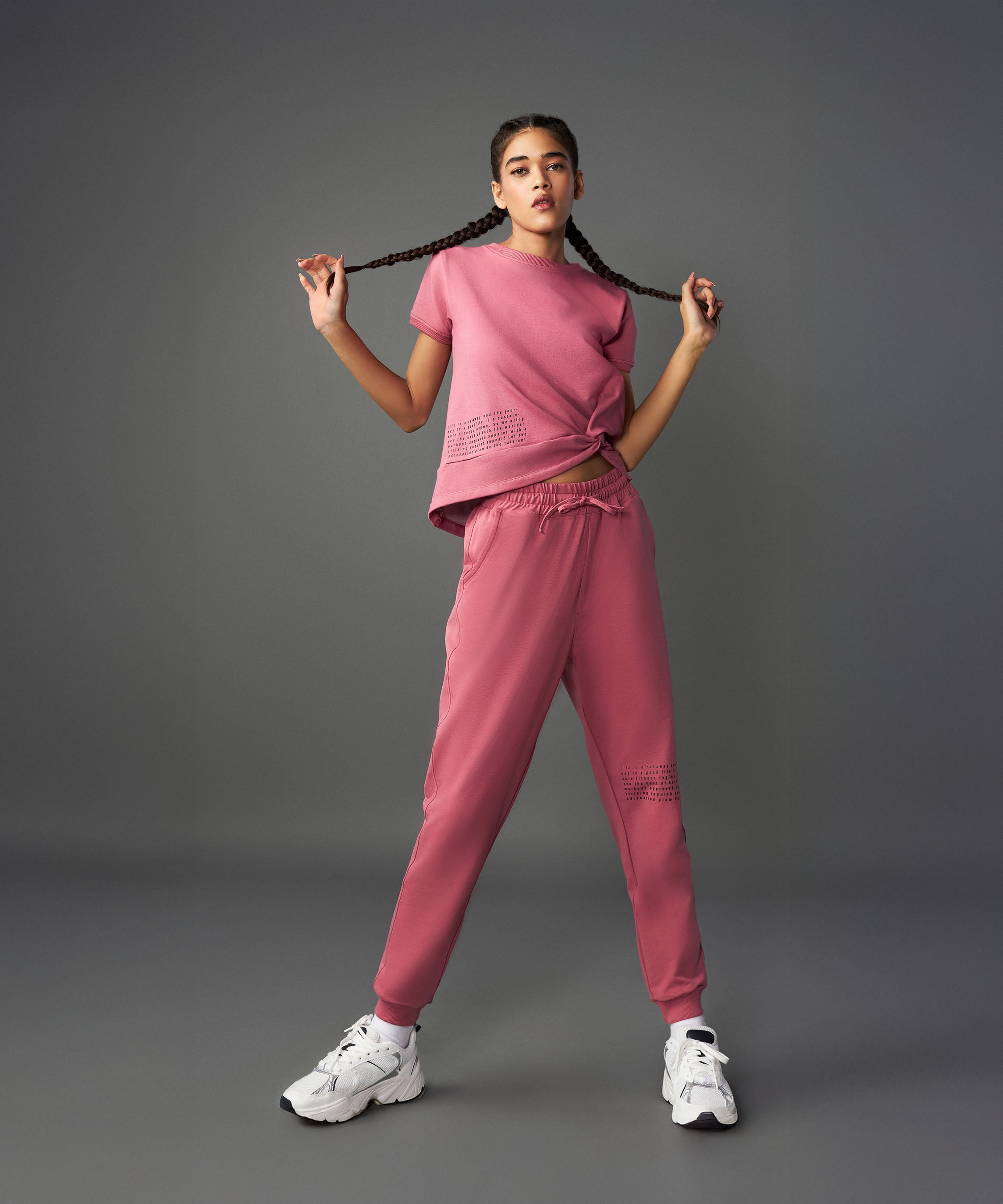 Woman wearing the Tuna Active Vintage Pink Co-ord Set with white sneakers, holding her braids in each hand against a gray background.