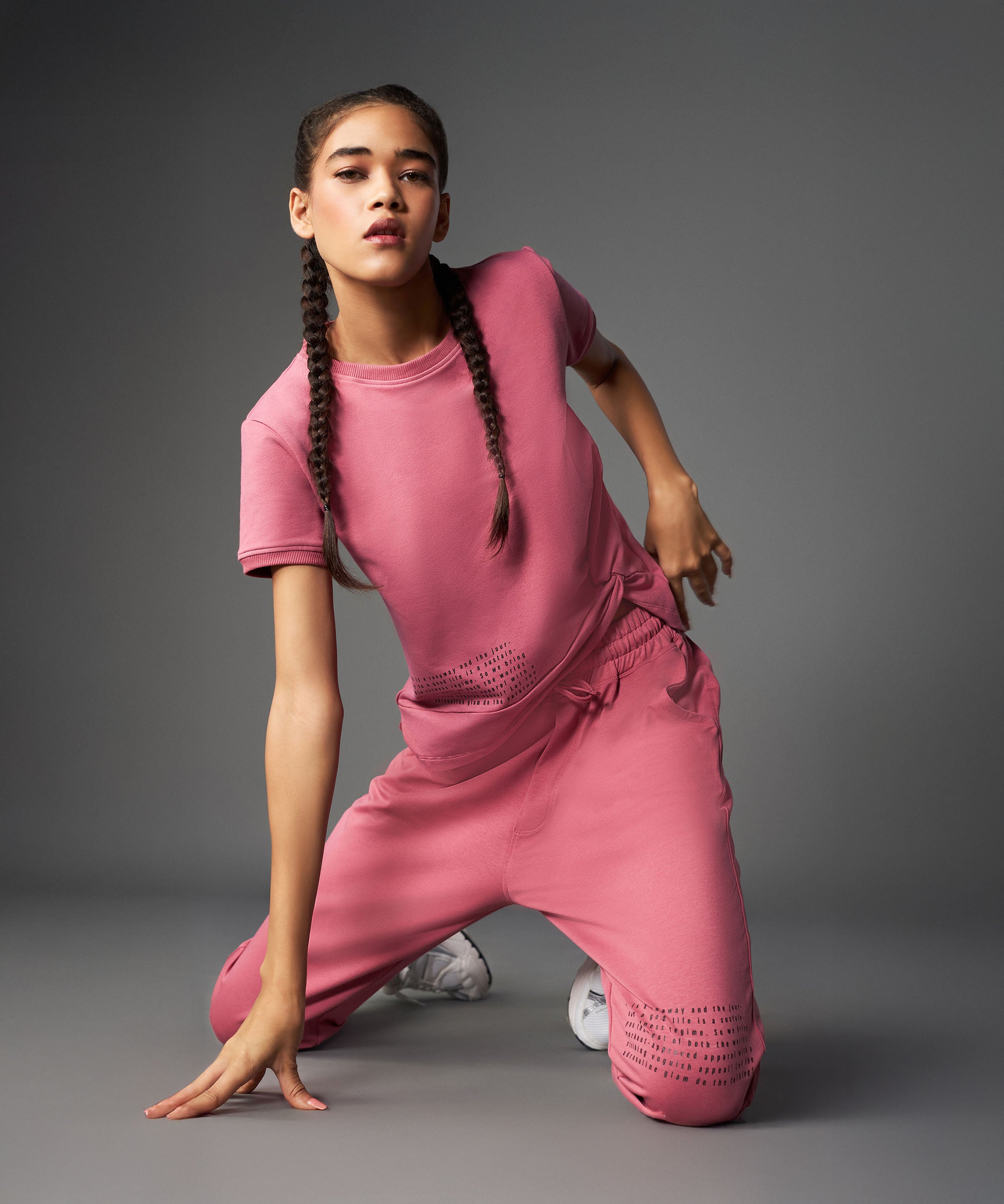 Woman wearing the Tuna Active Vintage Pink Co-ord Set kneels on one knee against a gray background, her hair styled in braids.