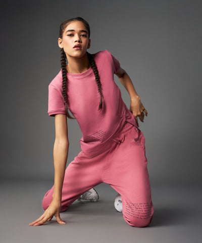 Woman wearing the Tuna Active Vintage Pink Co-ord Set kneels on one knee against a gray background, her hair styled in braids.
