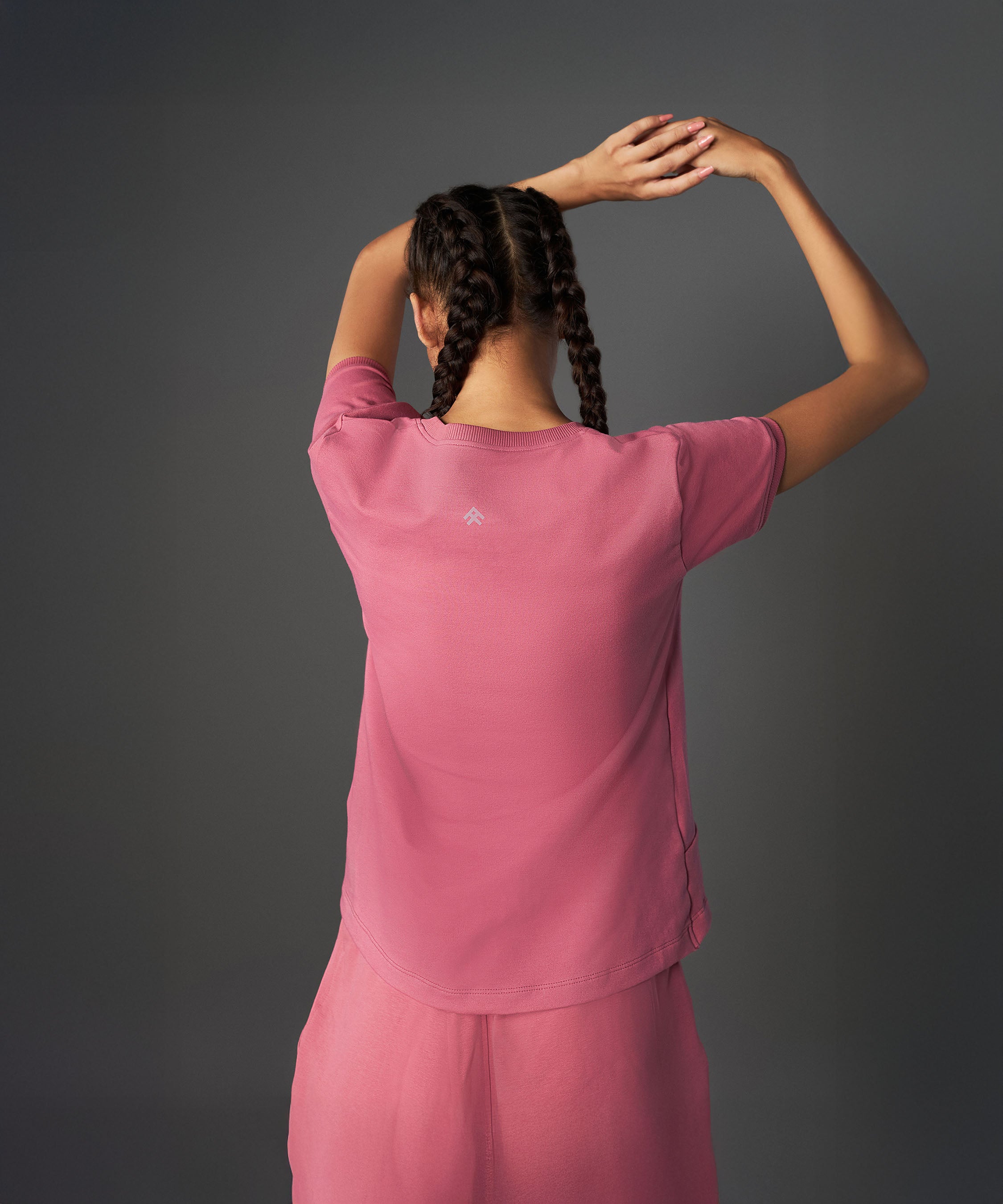 A woman with braided hair models the Tuna Active Vintage Pink Co-ord Set, facing away with arms raised overhead against a gray background.