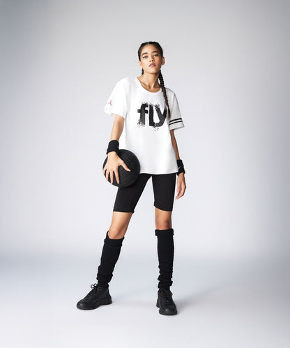 A young woman in a sporty outfit holds a basketball, wearing the Oversize Fly Tee with bold "FLY" print, black shorts, and long socks. The 100% cotton Oversize Fly Tee offers comfort both on and off the court.