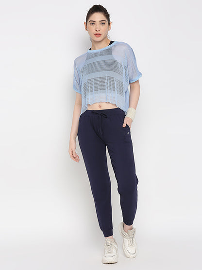 A woman stands against a white background wearing Blue Classic Joggers TLC04 with an adjustable fit, paired with a light blue sheer top and white sneakers—ideal for stylish lounge wear.
