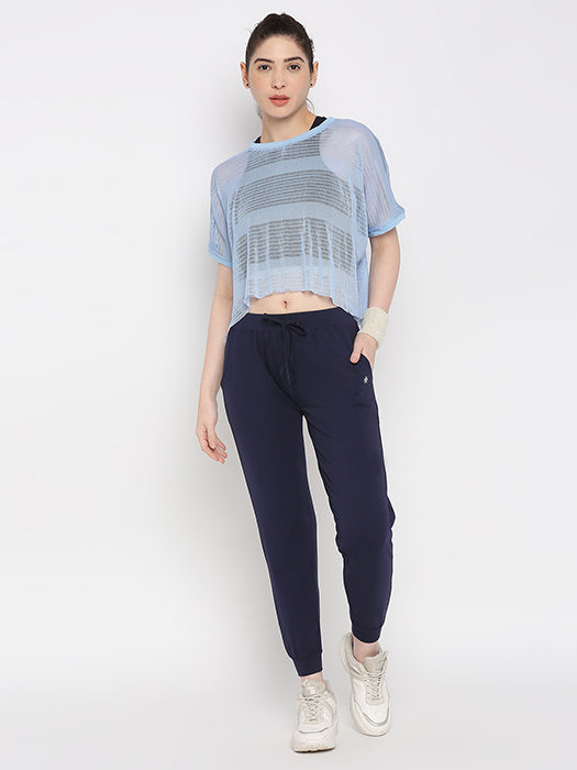 A woman stands against a white background wearing Blue Classic Joggers TLC04 with an adjustable fit, paired with a light blue sheer top and white sneakers—ideal for stylish lounge wear.