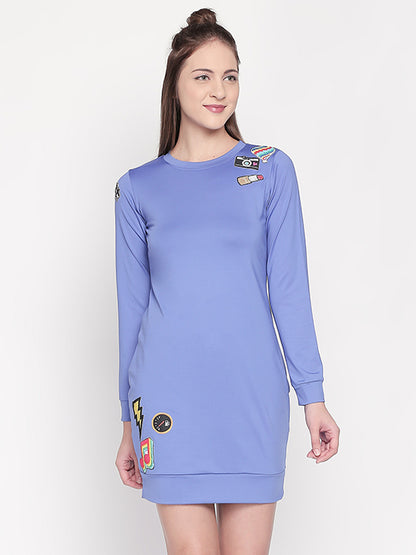 A woman wears the Cool Patched Full Sleeve Blue Dress by Tuna London, featuring colorful patches that create a playful fashion statement against a plain background.