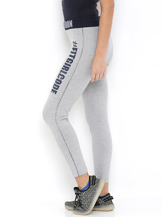 Woman wearing Narrow Fit Track Pants—high waist, grey, ankle-length—with "#FITGIRLCODE" text. Styled with black sneakers and a dark top, these pants offer supreme comfort and a trendy look while standing sideways.