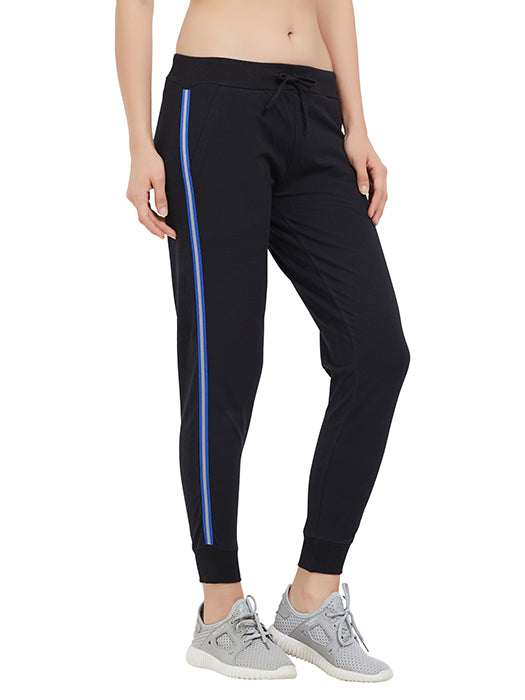 A person wears Classic Joggers TLC02SS in black with blue side stripes and a drawstring closure, paired with gray sneakers, standing against a white background.